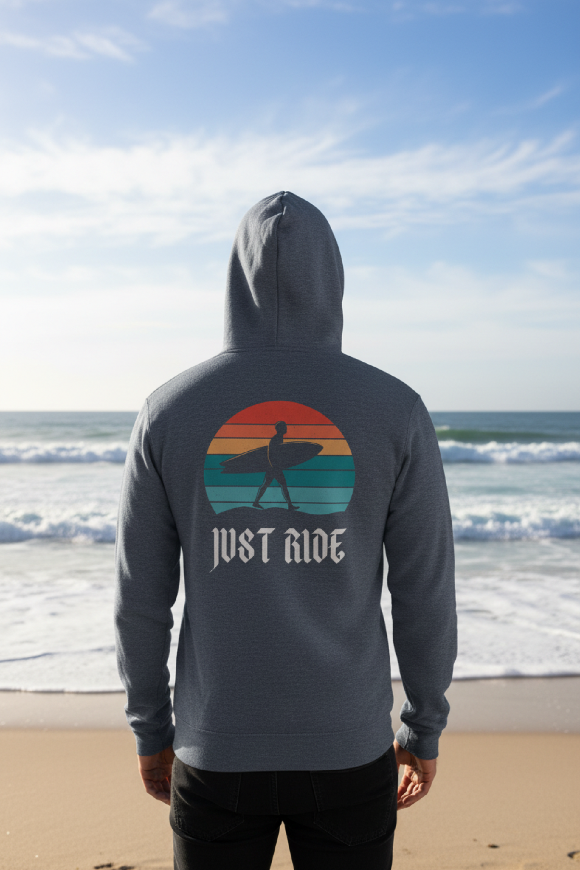 A person standing on a beach, viewed from behind, wearing a dark gray hoodie with a large retro sunset graphic and the words 'JUST RIDE' on the back. The background shows ocean waves, sand, and a blue sky.